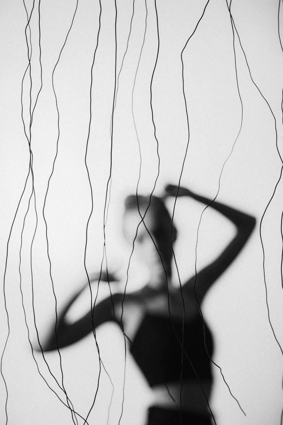 A woman's blurred silhouette, one arm raised behind her head, seen through hanging black wires on a pale screen.