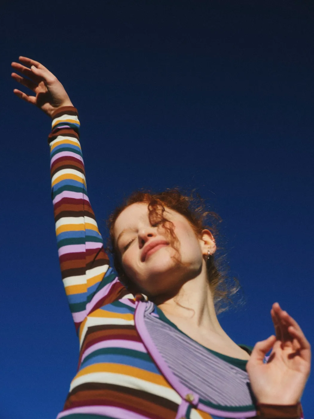 A woman in a striped top, one arm raised to a bright blue sky, eyes closed in the sun.