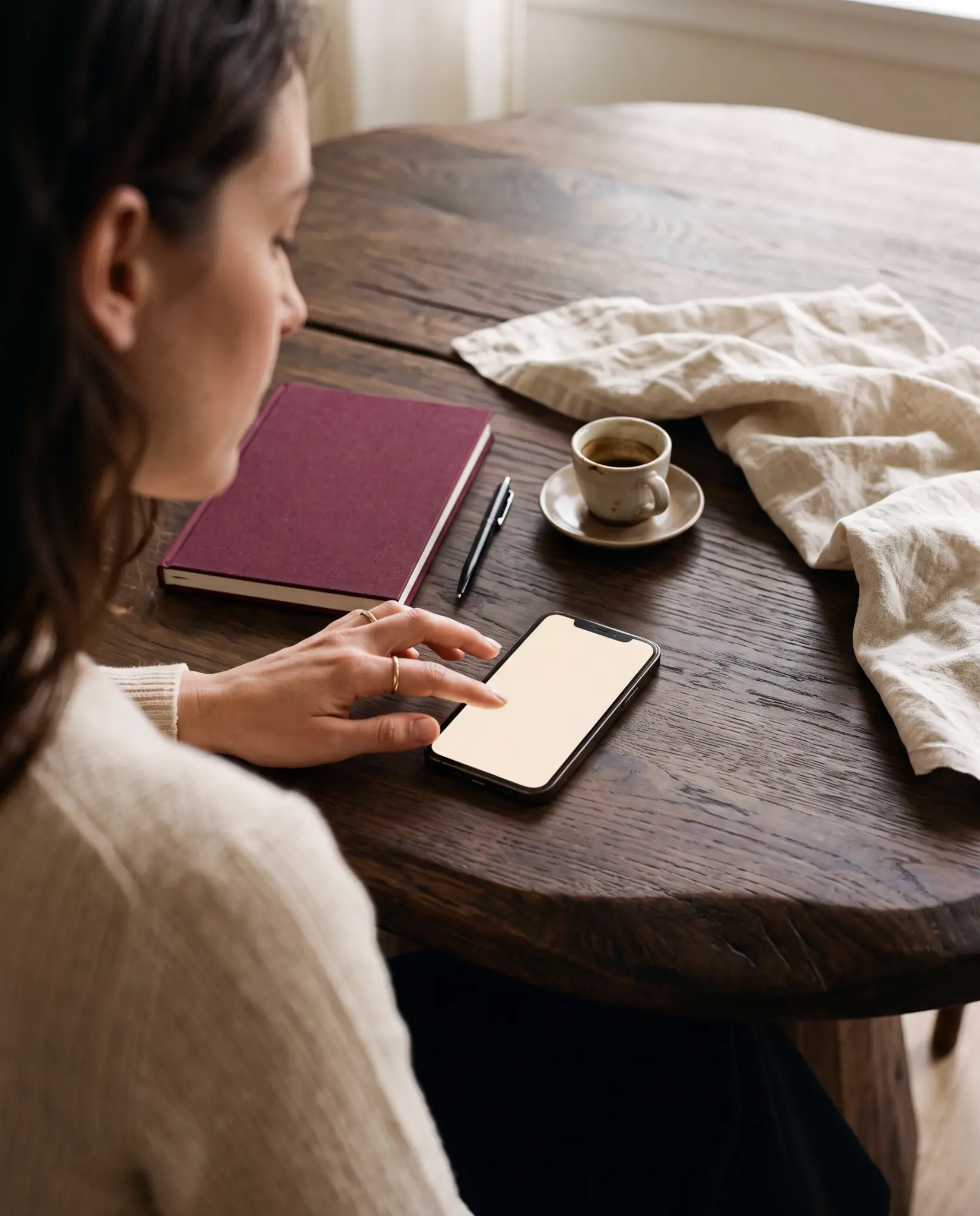 A hand on a phone at a wooden table beside a notebook and coffee